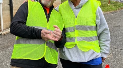 Two cheerful volunteers wearing bright yellow safety vests stand outside a brick building, serving cars for Packet Pickup at Lynchburg Turkey Trot. One is wearing a pink cap and sunglasses, while the other gestures playfully.